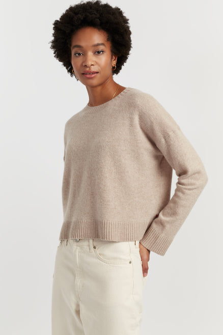 Woman wearing a beige sweater and white pants against a plain background